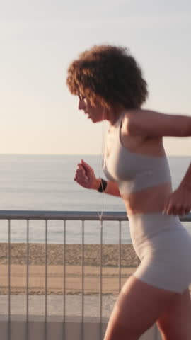 Vertical video, Woman runs along the beach at sunrise. She is wearing a light grey sports bra and shorts, and has earbuds in.
