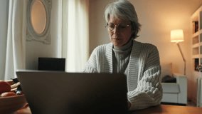 A senior woman with grey hair and glasses sits at a table and works from home on her laptop. She appears focused and sometimes thoughtful, and other times smiles. - Powered by Shutterstock - Get 15% off with code: PIKWIZARD15