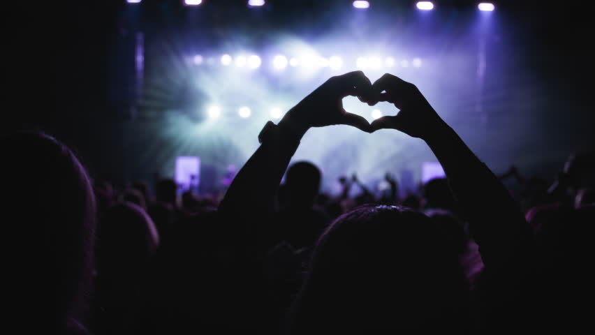 Concerthall show. Dark silhouette of female hands in the shape of heart fingers, blue backlight from the stage. Medium close-up shot. 