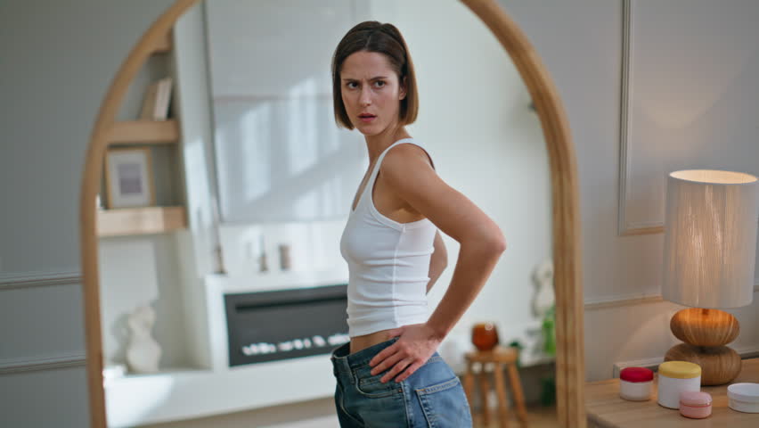 Sad lady trying jeans examining reflection in contemporary home interior. Slim woman dissatisfied weight loss looking mirror. Unhappy brunette wearing loose denim pants feeling displeased. Diet result