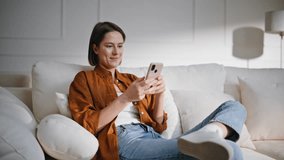 Home girl messaging smartphone sitting comfortably at soft couch pillows. Happy brunette laughing reading sms cellphone in cozy apartment. Smiling woman browsing phone social media relaxing at sofa. - Powered by Shutterstock - Get 15% off with code: PIKWIZARD15