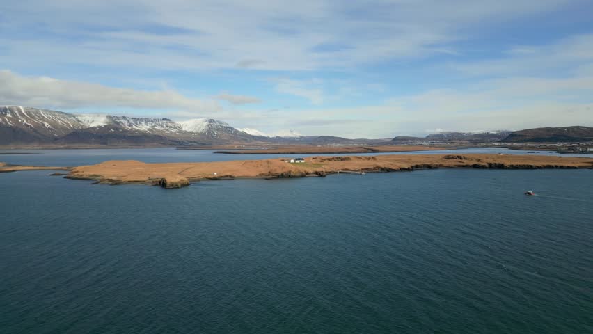 A scenic aerial view of a yellow lighthouse on the ocean with mountains in the background near Reykjavik, Iceland. A boat sails near a coastal house on the shore, creating a peaceful Nordic landscape.