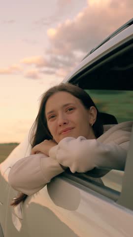 Teenage passenger riding in automobile. Cute girl is smiling from car window, wind is blowing her hair. Vacation trip. Young girl passenger is looking out window enjoying fresh breeze from car window