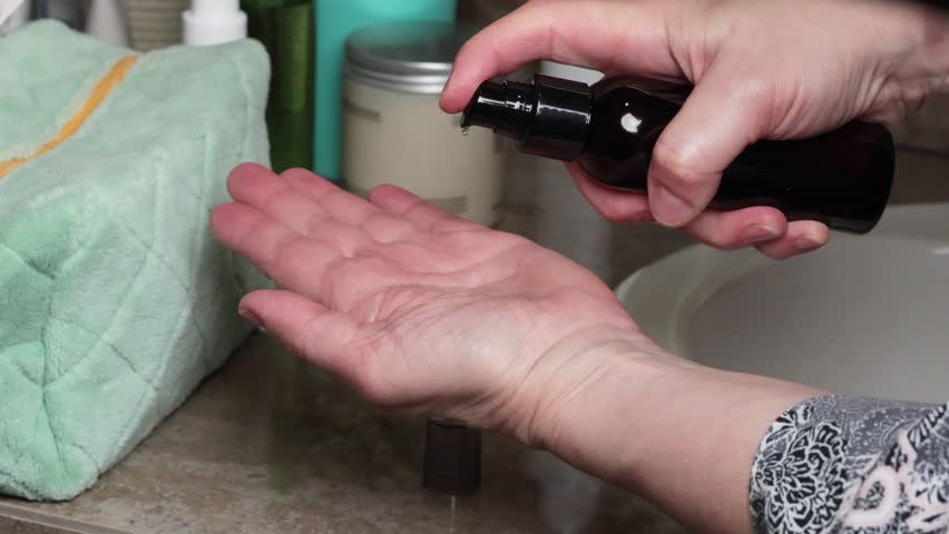 Woman in robe drips hair oil from bottle onto palm of hand. Beauty and Health. Lifestyle and self care. 