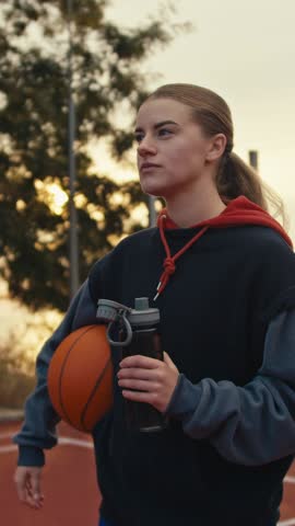 Focused blonde girl drinks water from a sports gray bottle while playing basketball on the street court