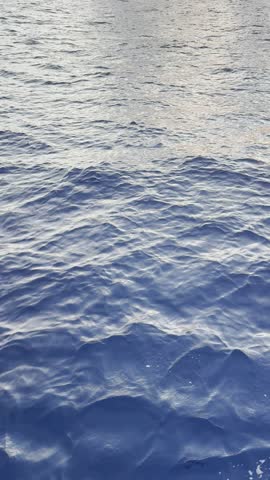 A serene slow motion scene of gentle ocean waves rippling across the water's surface. Sunlight shimmers on the ocean, creating a tranquil blend of blue and silver hues.