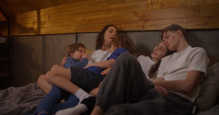 Happy brunette woman hugs her sons and lies on the bed with her teenage children at home in a cozy room