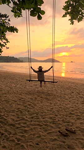 A serene moment on Koh Mak Island, where a person enjoys swinging gently while the sun sets over the tranquil waters. The golden hues reflect on the beach, enhancing the peaceful atmosphere.
