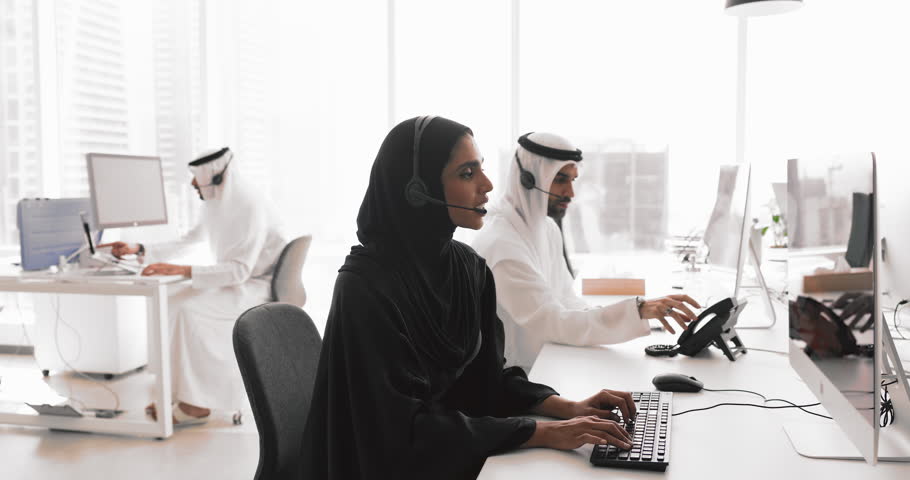 Group of Middle Eastern professionals dressed in traditional attires equipped with headsets sitting in coworking office, working in customer support or call center. Telemarketing, assistance services