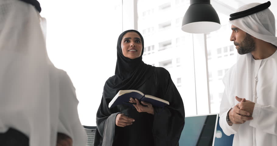 Leadership, empowerment, professional communication. Woman in black abaya holds notebook, speaks with confidence, share ideas, point and solutions with two male colleagues during collaborative meeting