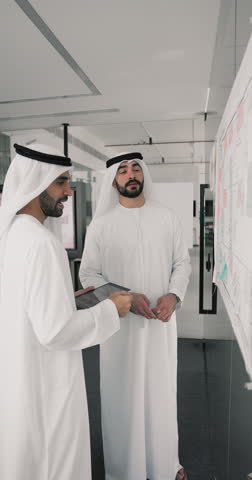 Two young Arabian colleagues in traditional white muslim dresses standing in office, review information attached on wall, discussing joint project updates, share ideas, creative solutions or deadlines
