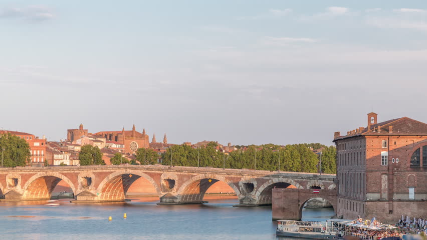 Garonne River and Pont Neuf timelapse with Museum of the History of Medicine in downtown Toulouse, France. This Renaissance arch bridge reflects in water under a blue sky. Waterfront with green trees