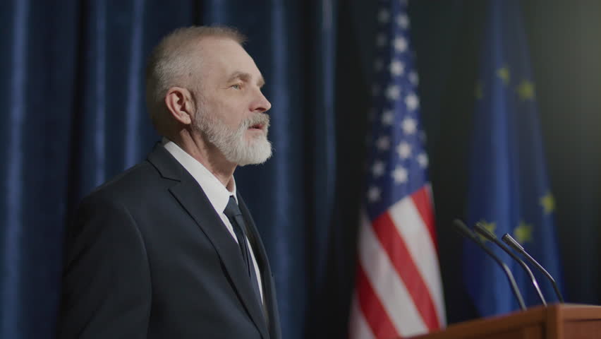 Senior male diplomat in formal suit standing on stage and answering questions from audience during international summit with American and European Union flags in background