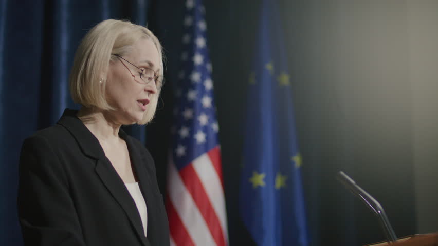 Middle-aged female diplomat delivering speech at podium with microphones during international summit with American and European Union flags in background