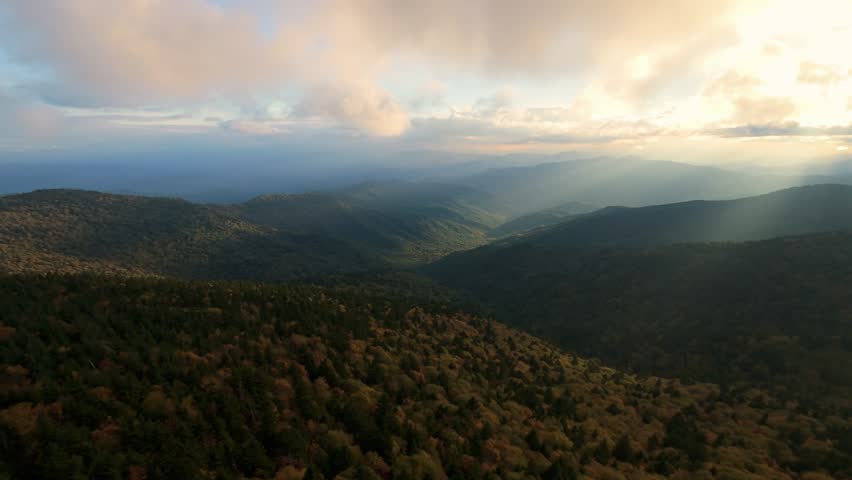 Golden light breaks through clouds, casting rays over vast forested mountain range in North America. Sun Rays Over Mountain Valley in USA or Canada drone flight forested mountain