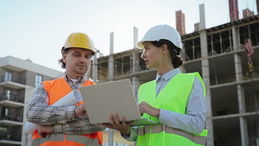 Male and female engineers in helmets and vests collaborating on laptop at construction site. Building framework in background of outdoor project discussion. Man and woman working as constructors.