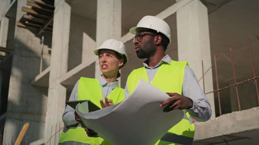 Multiethnic engineers discussing work at construction and making plan. Team of male and female working in safety helmets with building in background. Coworkers talking with drawing and tablet in hands