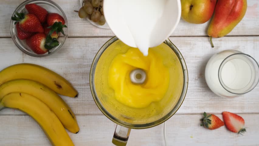 Top view pouring fresh milk into a mixed fruit yellow cocktail using a blender. Milk fruit drink obtained by chopping fruits and adding milk. Vegetarian food and diet. Morning fruit drink