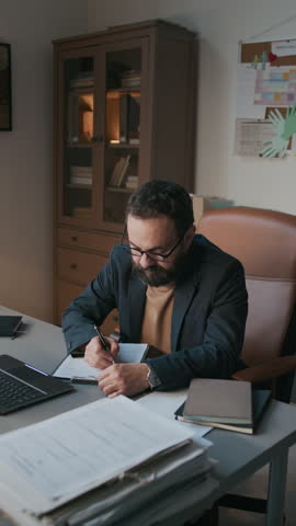 Vertical shot of male bearded psychologist in smart casual clothes and glasses sitting at desk and filling out mental health intake form after therapy session in modern office