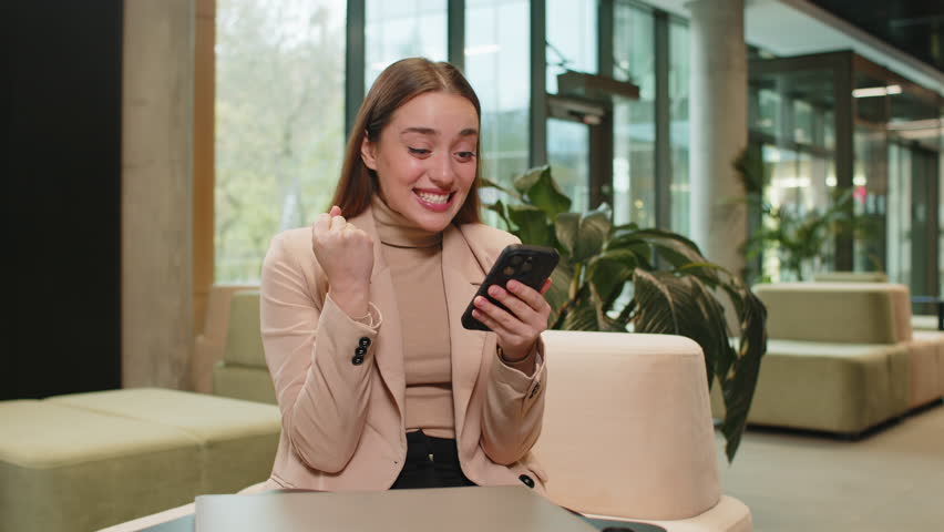 Happy businesswoman girl using mobile smartphone typing browsing, celebrating success victory, winning lottery casino jackpot achievement, play game, good positive news, triumph in modern office lobby