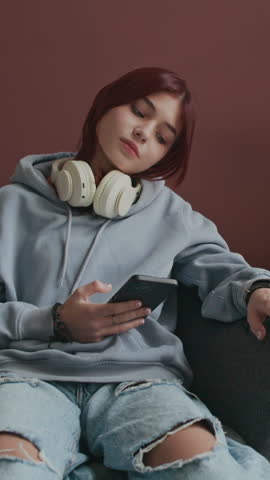 Vertical full shot of digital addict Caucasian teenager with red hair and headphones on neck staring at smartphone screen and showing lack of interest
