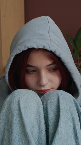 Vertical chest up shot of depressed teenage Caucasian girl in hood hugging knees while telling about bullying at school, she crying and covering head with hands, school pressure problems