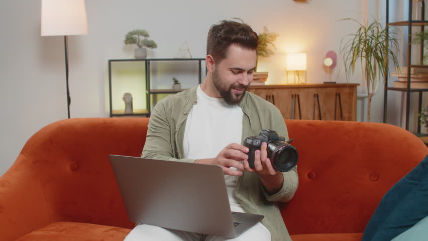 Portrait of happy young Caucasian brunette man photographer on sofa couch holding digital camera looking at screen, choosing photos for editing while sitting in front of laptop computer at home room.