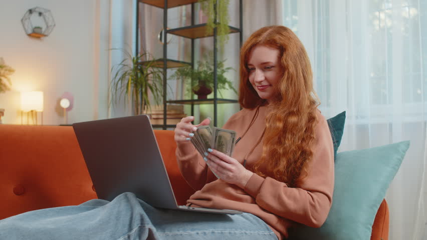 Planning budget. Rich happy young woman counting money dollar cash using laptop calculate domestic bills sitting on sofa. Girl satisfied of income earnings, saves money for planned vacation, gifts.