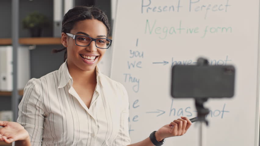 An African American woman teaches online, using a whiteboard to explain English grammar concepts. She interacts with students remotely, showcasing her expertise in education and tutoring.