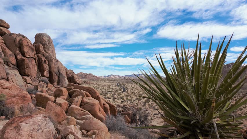 Slide motion to the left to reveal the main road into Joshua Tree National Park from atop a small mountain of boulders. Partly cloudy skies and expansive views into the distance.