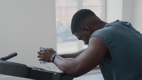 Tired Black man leaning on treadmill, drinking water and wiping sweat from forehead while recovering after cardio workout in home gym - Powered by Shutterstock - Get 15% off with code: PIKWIZARD15