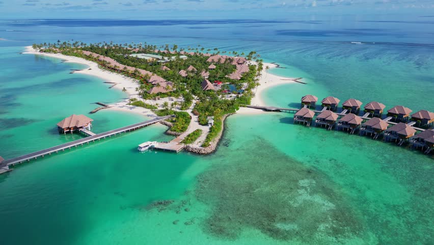Aerial rising shot of tropical island in Maldives during sunny day. Luxury huts and apartments surrounded by coral reef in ocean. Wide shot.