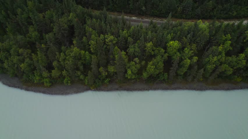 Aerial top down rising shot of natural lake and green trees with railway tracks in Alaska USA.