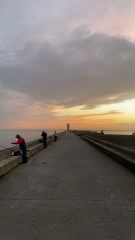 People enjoy fishing on a pier at sunset, casting lines into the ocean as the sky glows with warm colors in this tranquil coastal scene.