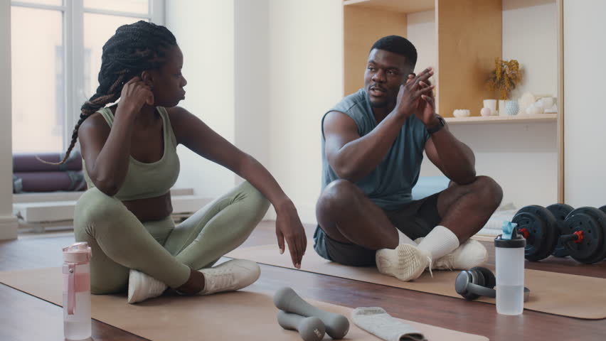 Young Black couple in sportswear sitting cross-legged on exercise mats, drinking water and chatting during break in home gym