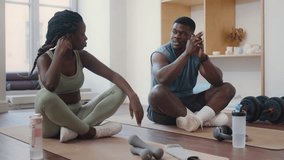 Young Black couple in sportswear sitting cross-legged on exercise mats, drinking water and chatting during break in home gym - Powered by Shutterstock - Get 15% off with code: PIKWIZARD15