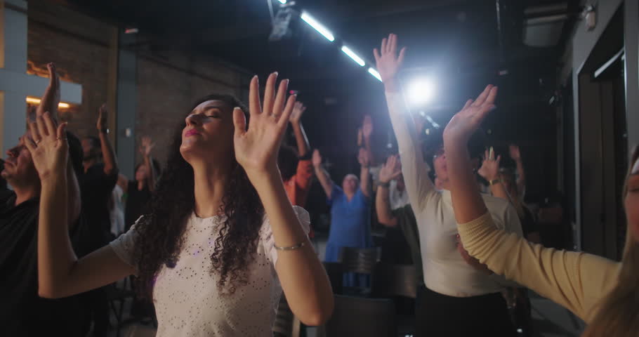 Worshipers raising hands in spiritual praise, diverse group with a man in focus, modern church background, illuminated cross in background