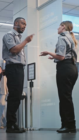 Crowded International Airport Terminal: Two Diverse Security Officers Talking, Standing at Security Checkpoint, Helping Woman. Diverse People with Luggage Going for Boarding Flights. Vertical Shot.