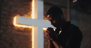Man with head bowed in prayer, holding hands together near illuminated cross, deep spiritual reflection, faith and devotion, Christian worship, contemplative moment - Powered by Shutterstock - Get 15% off with code: PIKWIZARD15