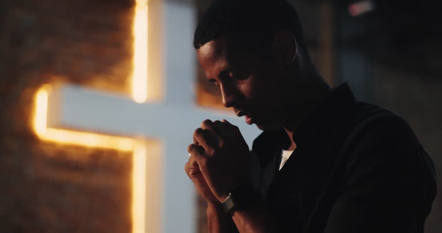 Man with head bowed in prayer, holding hands together near illuminated cross, deep spiritual reflection, faith and devotion, Christian worship, contemplative moment