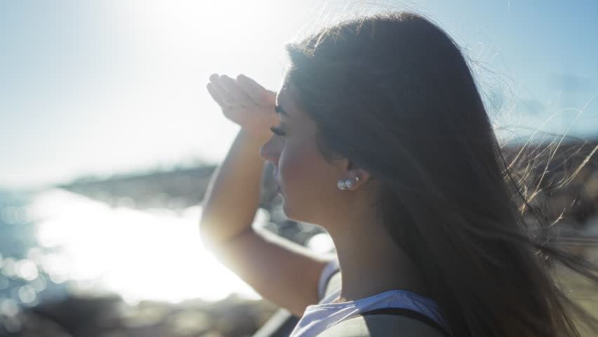 Woman enjoying sunny seaside promenade, shielding eyes from bright sun, with windblown hair and ocean background.