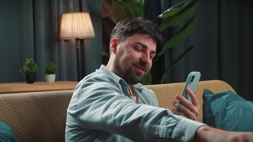 Junk food advertisement concept. Bearded man spending time at home with fast food delivery, sits on couch watching sport game on smartphone and eating chips, happy wining expression.