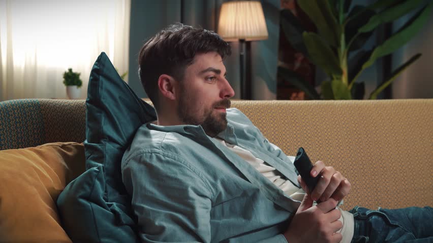 Junk food advertisement concept. Bearded man spending time at home with fast food delivery, lays on couch, watching tv eating potato chips and scrolling smartphone.