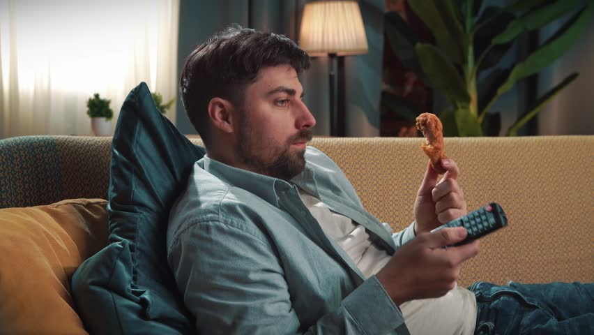 Junk food advertisement concept. Bearded man spending time at home with fast food delivery, lays on couch, watching tv switching channels and eating fried chicken.