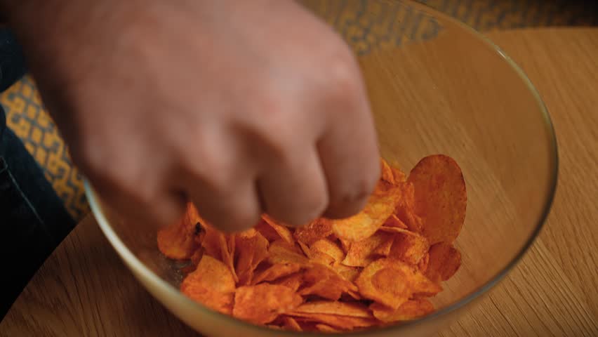 Junk food advertisement concept. Close up shot of bearded man sitting on couch, man taking and eating potato chips from a glass bowl, enjoying eating fast food. Slow motion