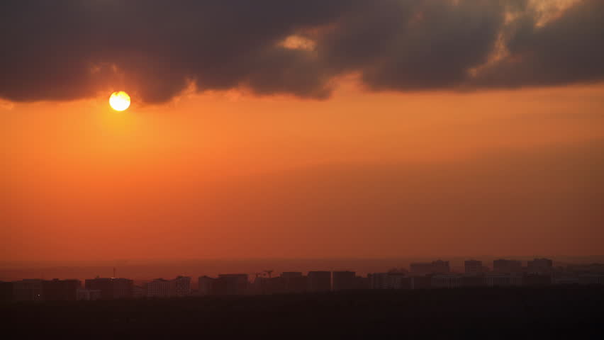 Sunset over city skyline with dark clouds, time lapse. Urban landscape with orange sky and setting sun. Cityscape with copy space.