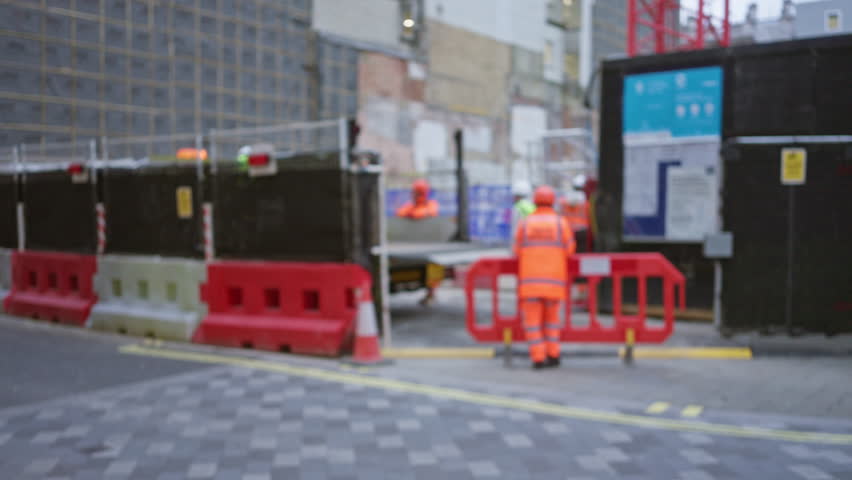 Blurred construction workers on site with orange uniforms and safety equipment by red barriers on street with defocused urban background