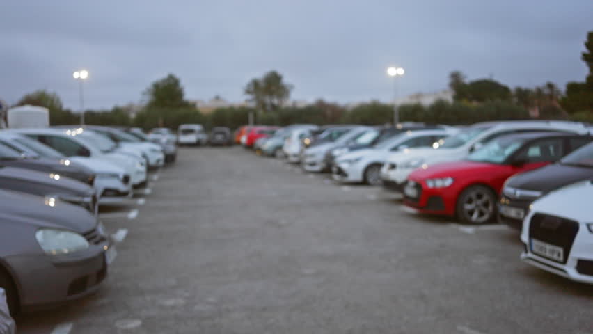 Blurred view of a parking lot with numerous cars lined up, showcasing a defocused urban setting under cloudy skies with scattered streetlights creating a serene atmosphere.