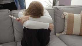 Woman sitting on a sofa with arms around knees holding a pregnancy test, while a man stands near her in a modern living room, conveying emotion and relationship dynamics. - Powered by Shutterstock - Get 15% off with code: PIKWIZARD15