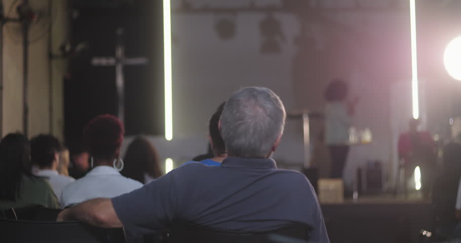 Group of people attending a church event, with a man in the foreground seated, and a woman speaking at the podium in a modern church with a cross in the background